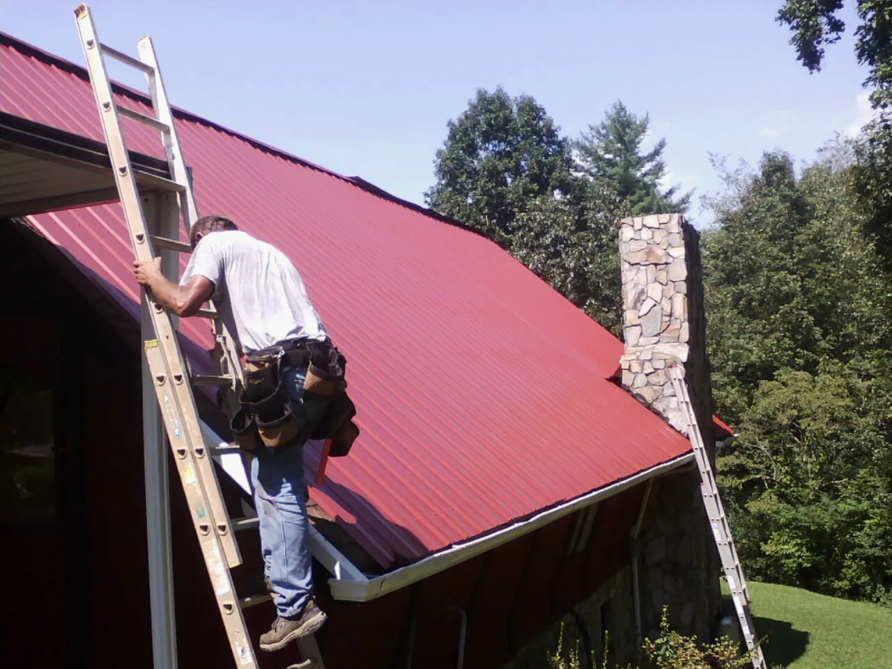 Roofer on a ladder beside a red metal roof and stone chimney — Soffit & Fascia Repair by Roofing Brothers in Landenberg